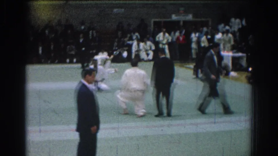 1966 San Gabriel gym competition with suited officials in the foreground and martial arts competitors active on the mat.