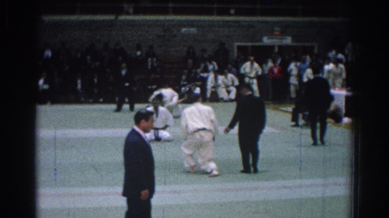 Two judo competitors grapple on a green mat while a dark-suited referee stands nearby.