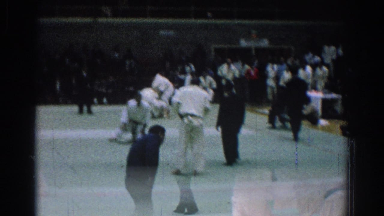 Judo competitors continue grappling on the mat with a referee and spectators in the gym.