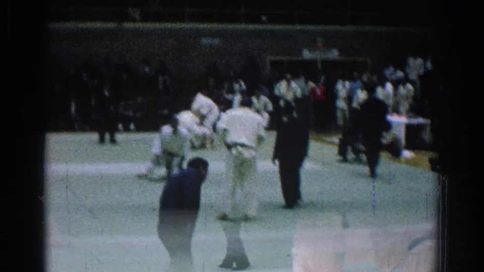 1966 San Gabriel judo match frame with athletes in white gi, a dark-clothed referee, and spectators seated along the back of the gym.
