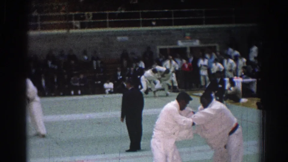 Home-movie frame from a 1966 San Gabriel gym showing judo athletes gripping each other, with a referee and crowded bleachers in view.