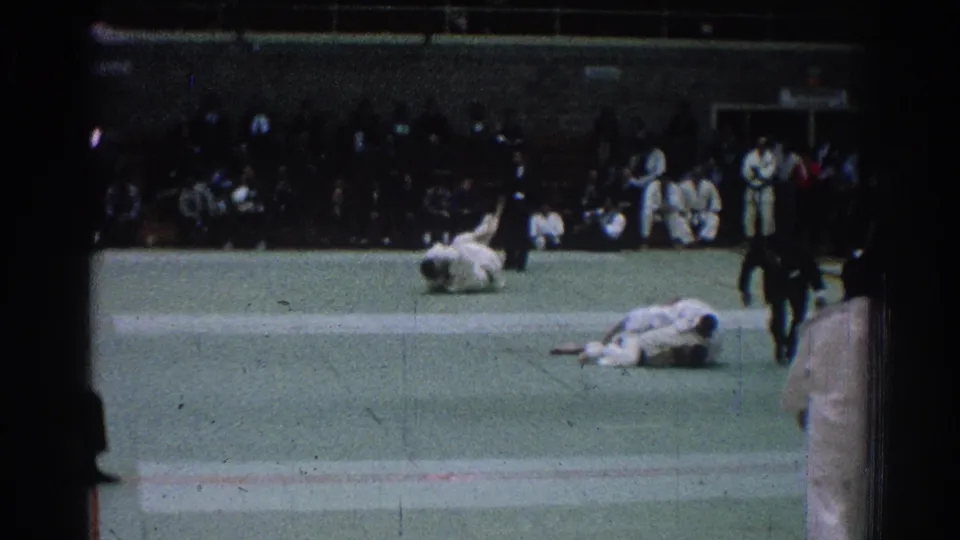 San Gabriel 1966 judo competition frame showing athletes on the mat, a referee standing nearby, and spectators seated along the gym wall.