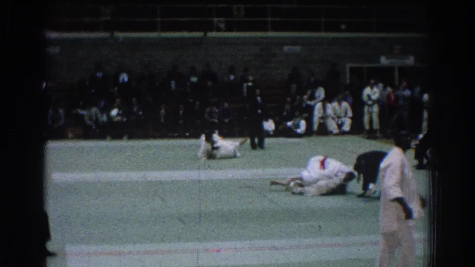 1966 San Gabriel judo match with competitors in white gi on a green mat, a referee nearby, and spectators seated along the back wall.