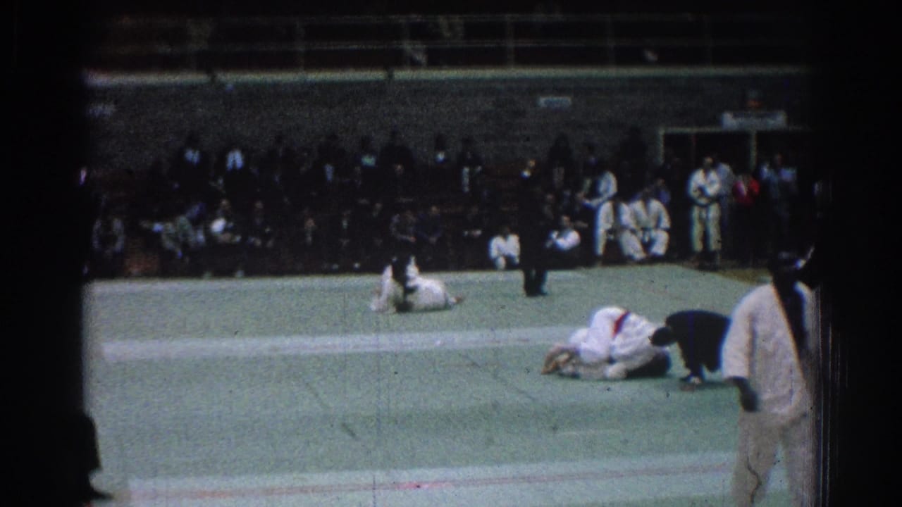 Two judo competitors are down on the mat with spectators seated behind them.
