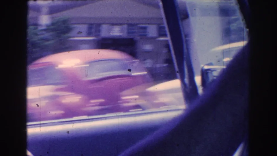 A red car blurs past the camera near the car window frame, with other vehicles visible on the street.