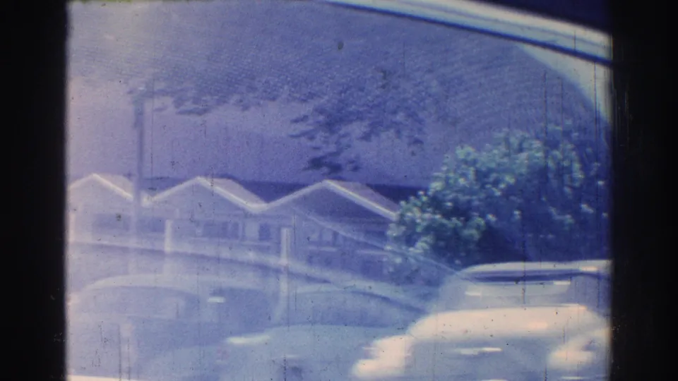 Blue-tinted 8mm view of San Gabriel residential facades and passing cars, partially obscured by a tree and vehicle frame.