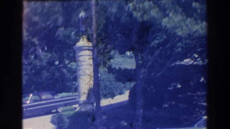 San Gabriel park scene showing a pale stone monument or marker among trees, with a roadway and landscaped edge nearby.