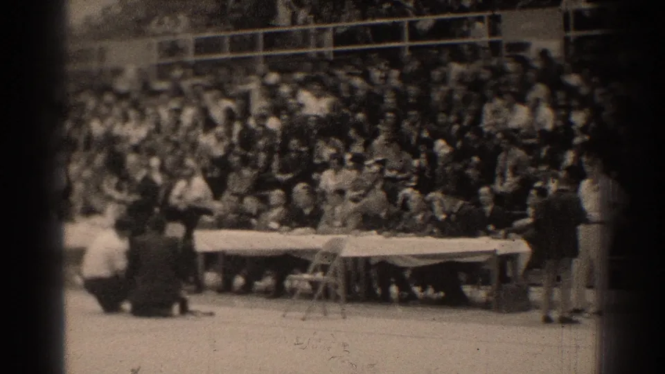 Outdoor gathering in San Gabriel with spectators seated in bleachers, a long table in front, and several people standing nearby.