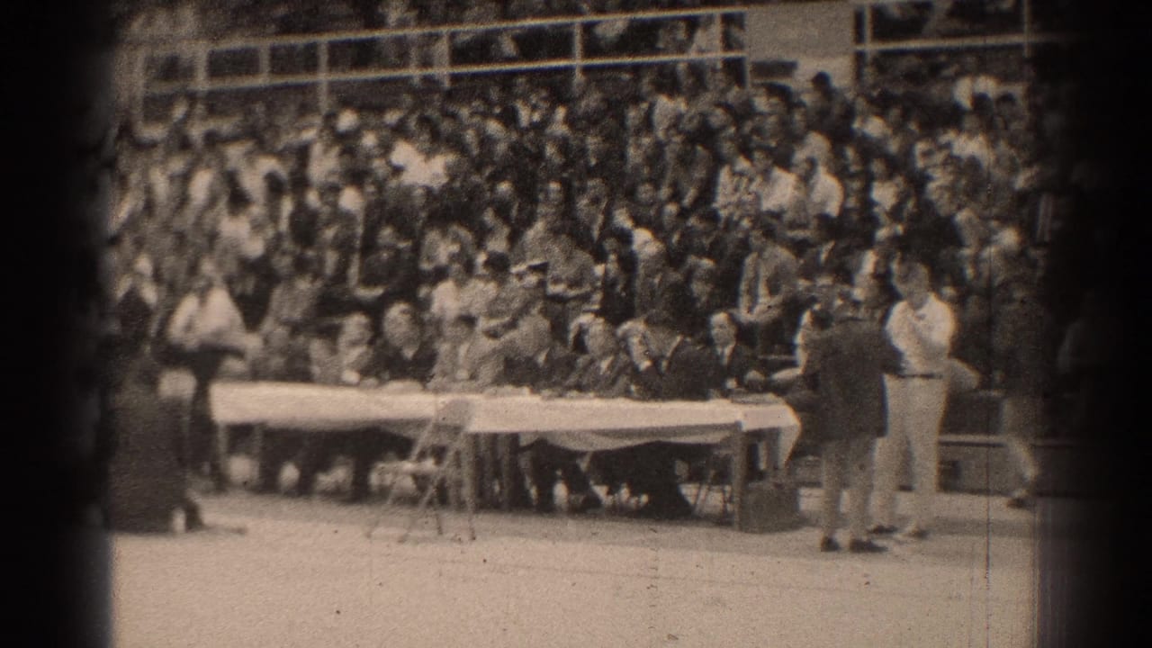 Crowded bleachers face a long white table during an indoor formal gathering.