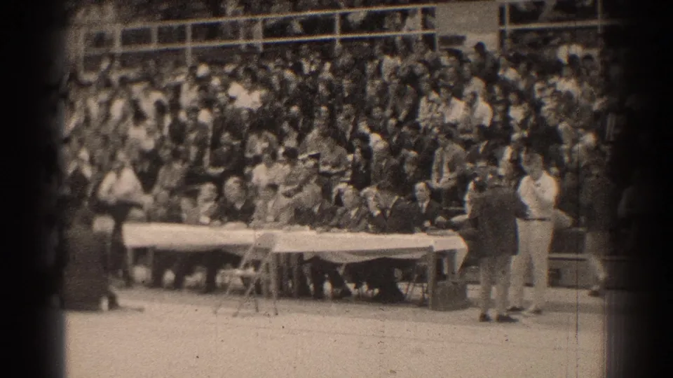 1966 San Gabriel home-movie frame showing spectators in bleachers watching a long white-draped table in an indoor arena.