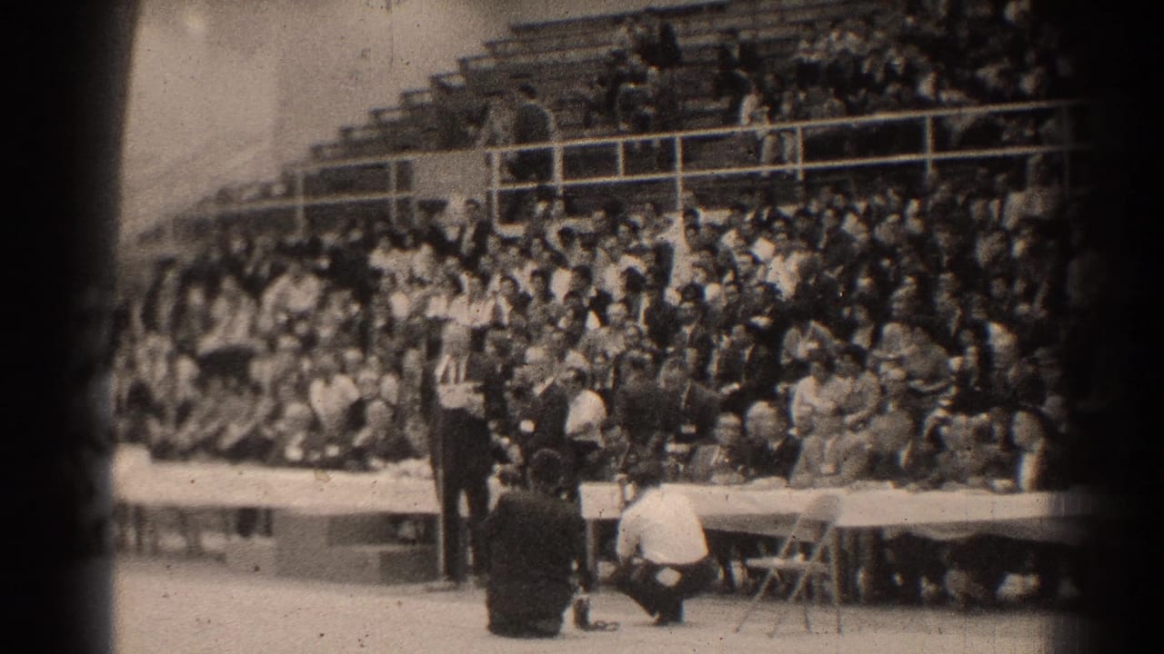 A speaker area and long table sit in front of crowded indoor bleachers.