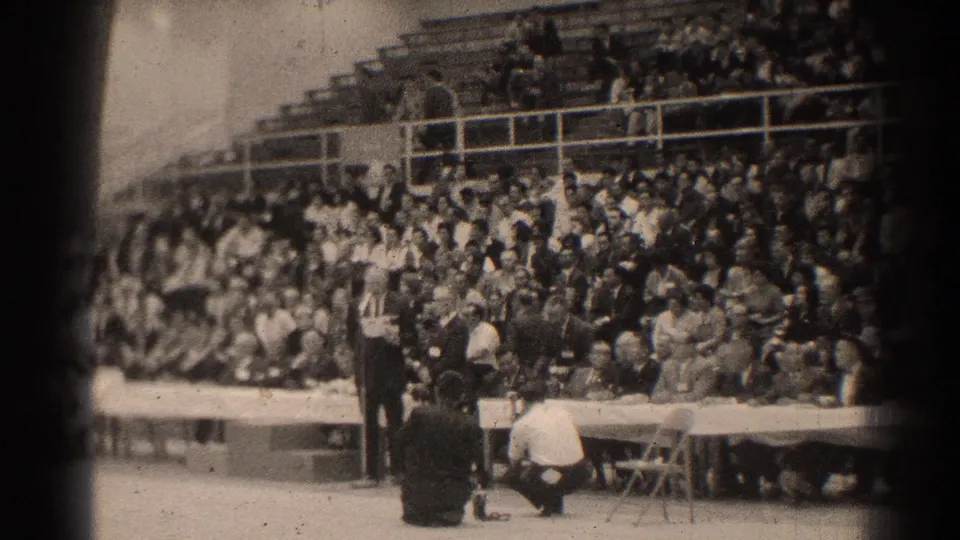 1966 San Gabriel frame with a standing figure near a microphone area, a long table, and spectators seated in bleachers.