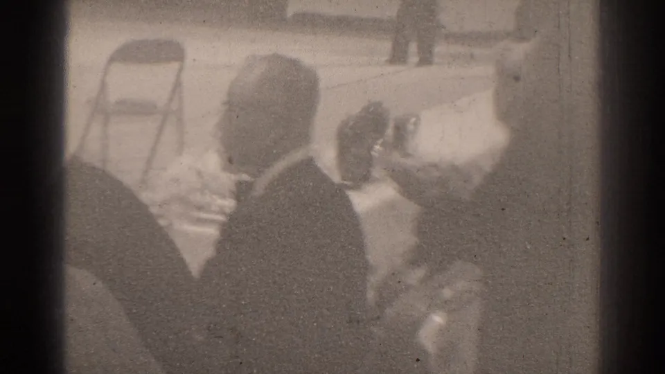 Soft 8mm frame from the San Gabriel gathering, showing seated figures near the table with the arena floor beyond.