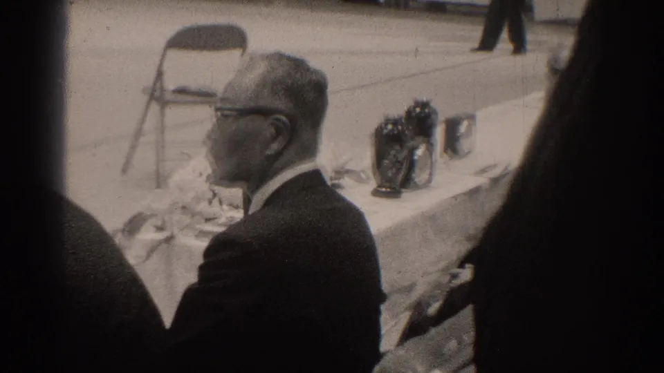 1966 San Gabriel frame showing a suited man with glasses beside a white-draped table and trophy-like objects during the event.