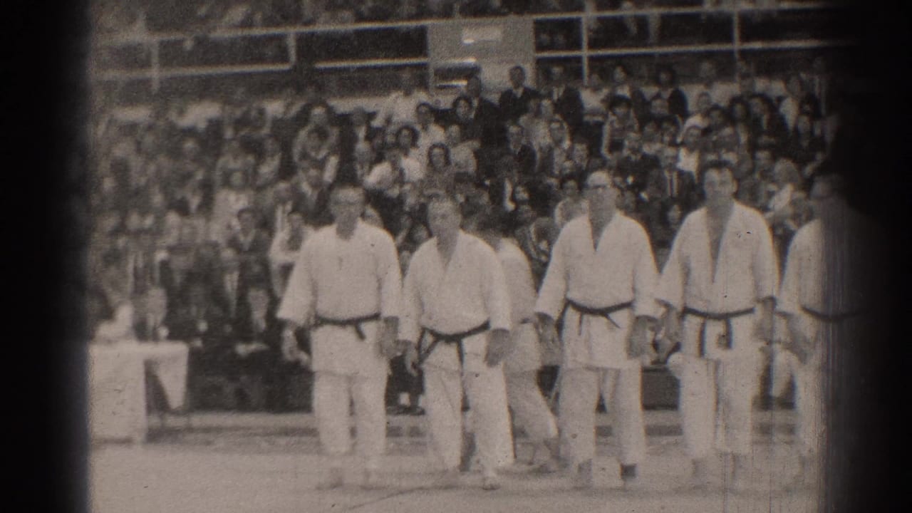 Men in white karate uniforms stand in line on a gymnasium floor before seated spectators.