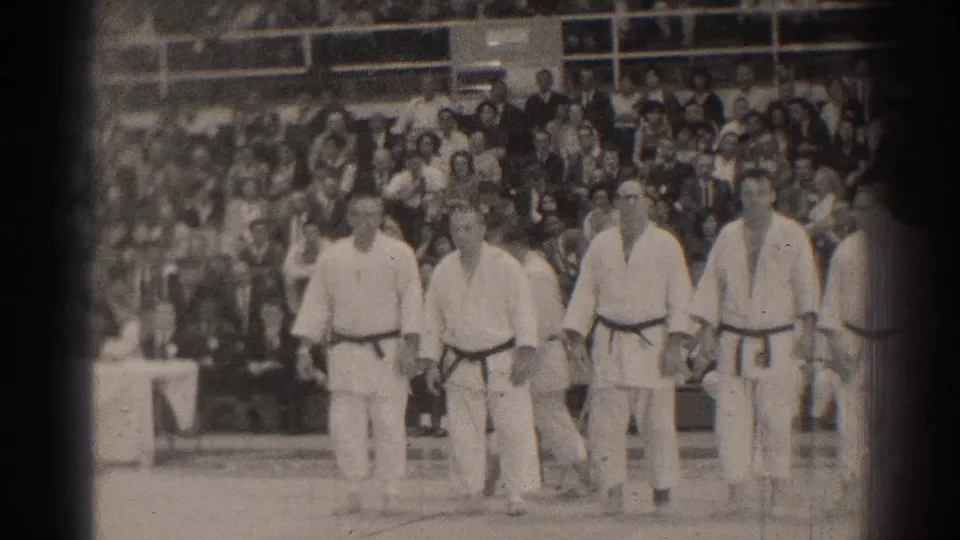In 1966 San Gabriel, men in white karate uniforms and dark belts stand before a large audience in bleachers.