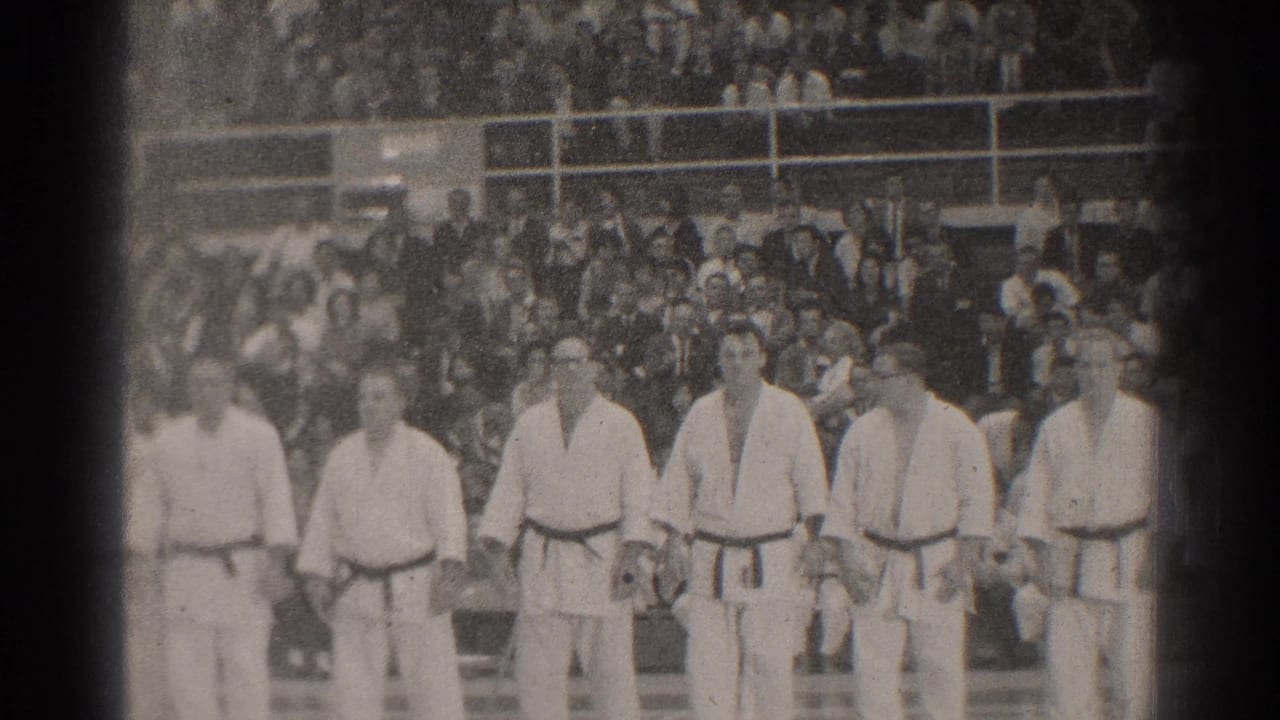 A line of karate practitioners faces forward with spectators seated behind them.