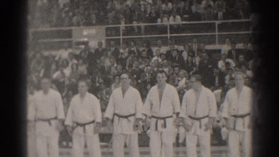 Men in karate uniforms stand in formation inside a San Gabriel gymnasium, with bleachers full of viewers behind them.