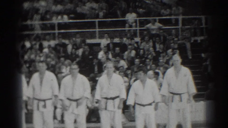 The final frame keeps the lineup of men in white karate uniforms before a large indoor audience in San Gabriel.