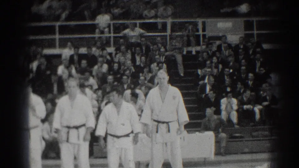 Several judo athletes stand in formation on a mat during a 1966 San Gabriel, California event, with spectators seated behind them.
