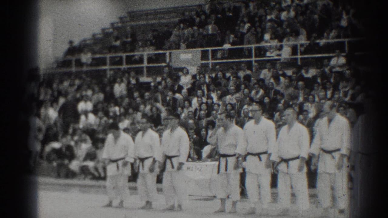 A row of judo athletes stands at the edge of the mat with bleachers behind.