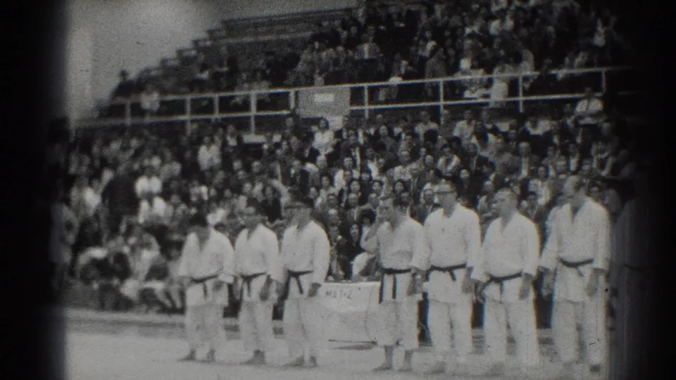 Judo competitors in white gi and dark belts stand in a row at a 1966 San Gabriel, California competition, with a packed audience behind them.
