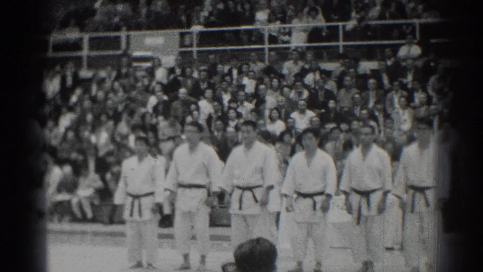 Judo athletes stand across the mat during a 1966 San Gabriel, California event, framed by black-and-white 8mm film edges and a large audience.