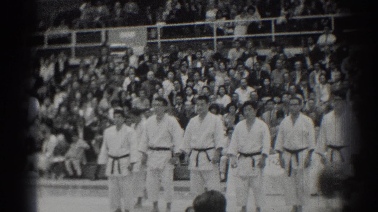 Martial artists in white gi stand in a line before crowded bleachers.
