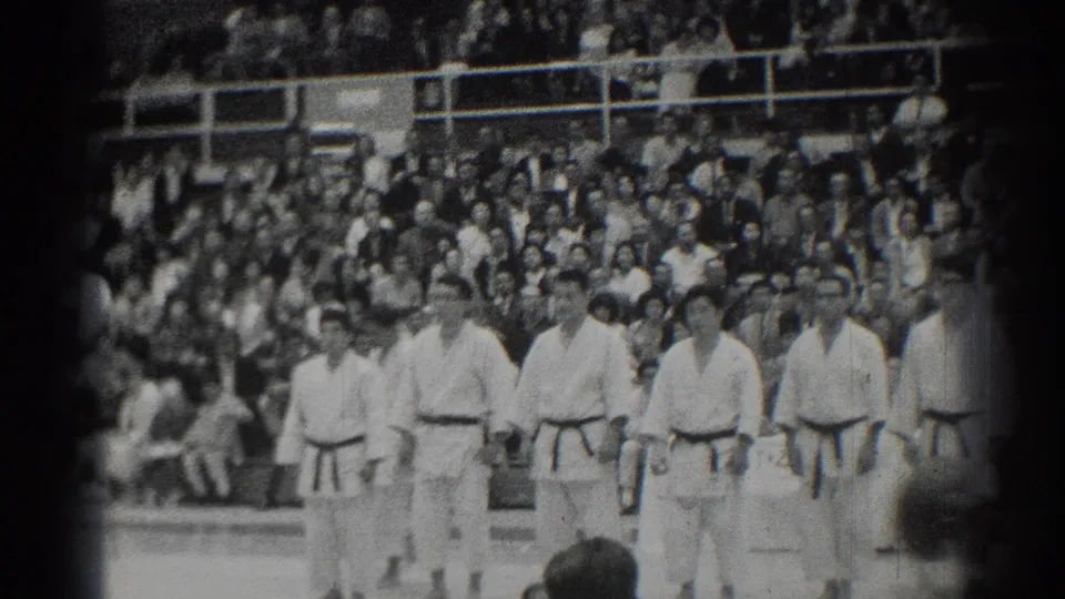 In a 1966 San Gabriel indoor arena scene, martial artists in white gi and dark belts stand at attention before spectators.