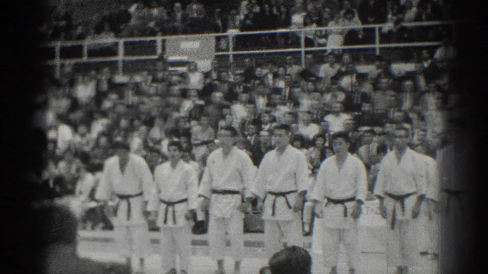 A black-and-white 8mm frame shows a formal martial arts lineup in San Gabriel with a dense seated audience behind.