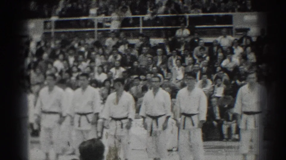 The home-movie frame shows a suited official or presenter near the gi-clad lineup during the 1966 San Gabriel martial arts presentation.