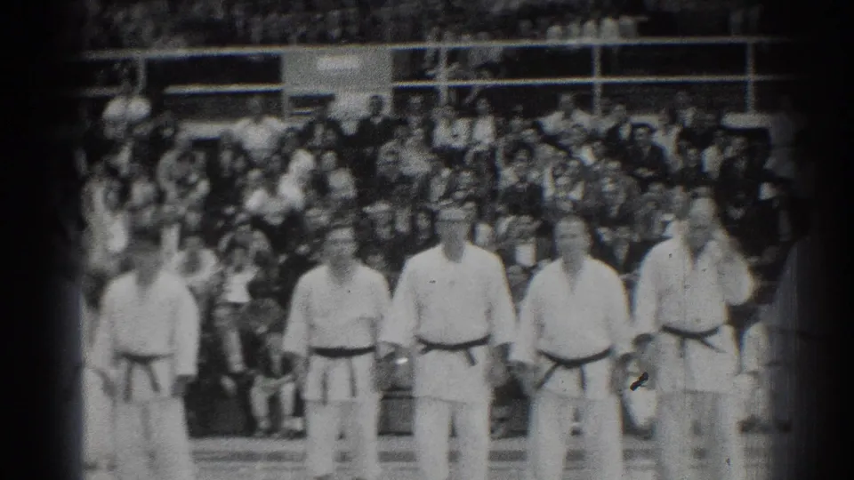 A soft black-and-white frame shows gi-clad martial artists lined up at an indoor San Gabriel event with spectators behind them.