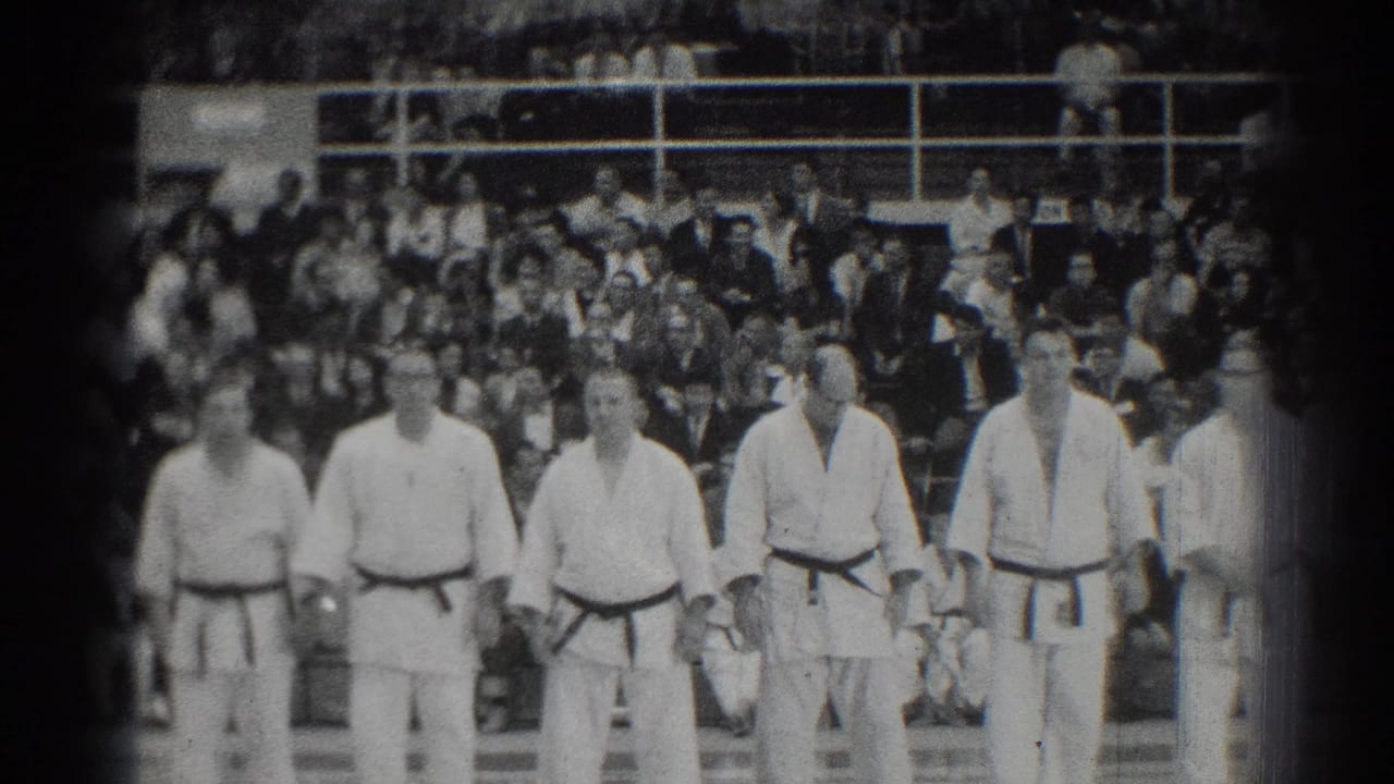 Judokas in white gi stand in a line before gym bleachers filled with spectators.