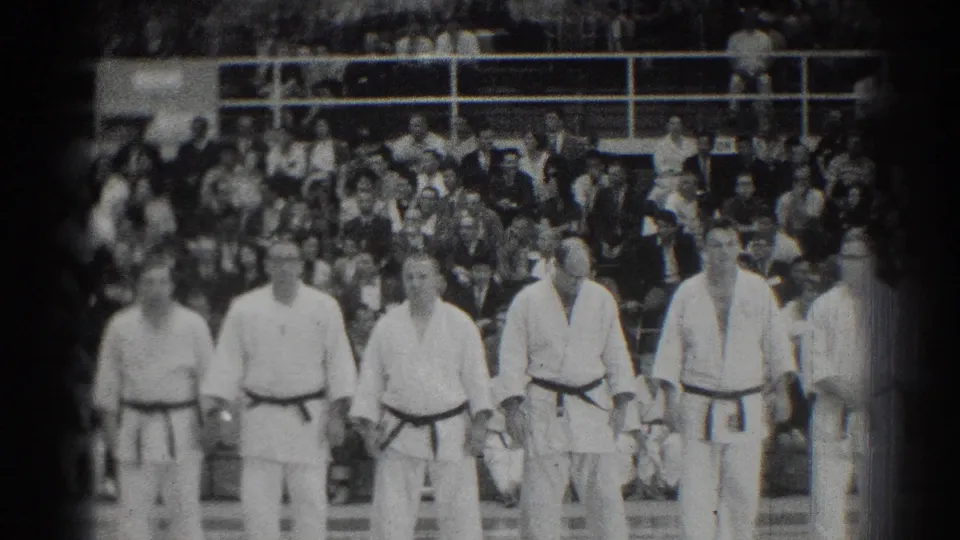 1966 San Gabriel judo competitors in white gi and dark belts stand at attention before a seated gym audience.