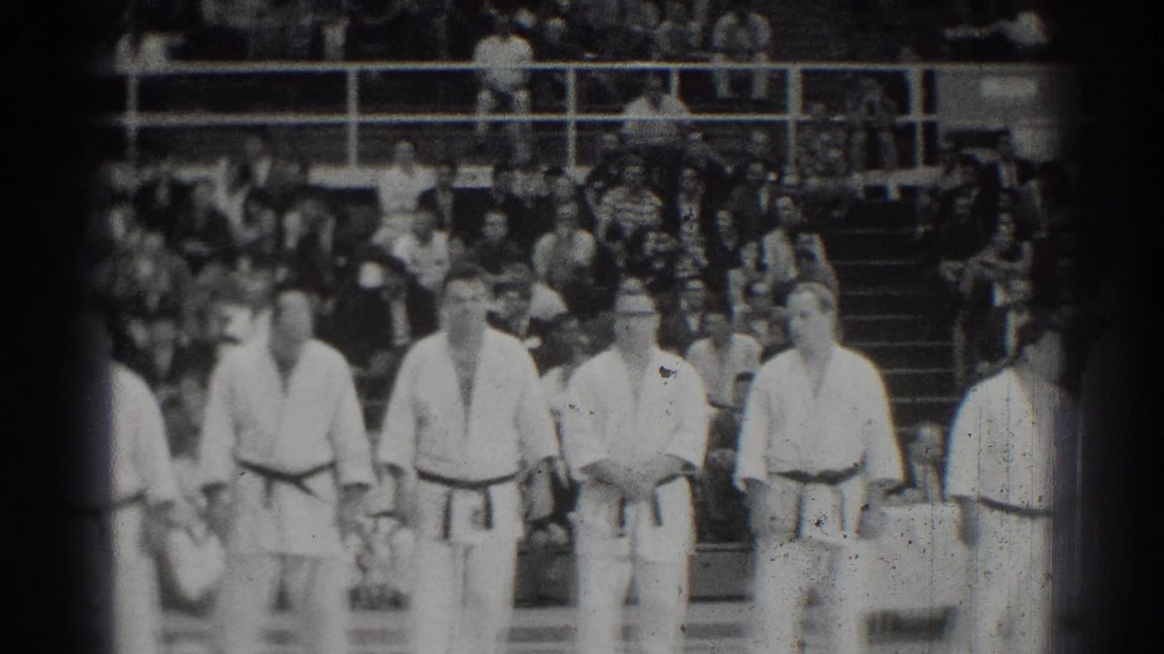 Several judokas stand in front of crowded bleachers, with one official-like figure near the line.