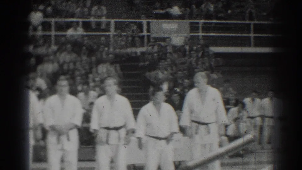 Soft 8mm frame of 1966 judo participants standing together in a gymnasium with an audience behind them.