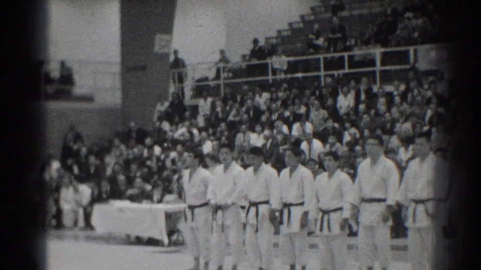 Wide 1966 San Gabriel gymnasium view with judo competitors lined up near a table before a large audience.