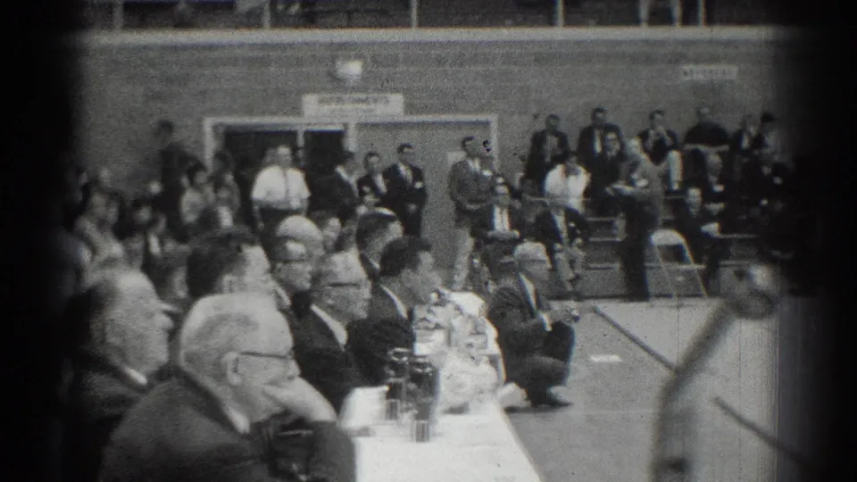 1966 home-movie frame from San Gabriel showing officials at a table and spectators watching a karate event in a gymnasium.