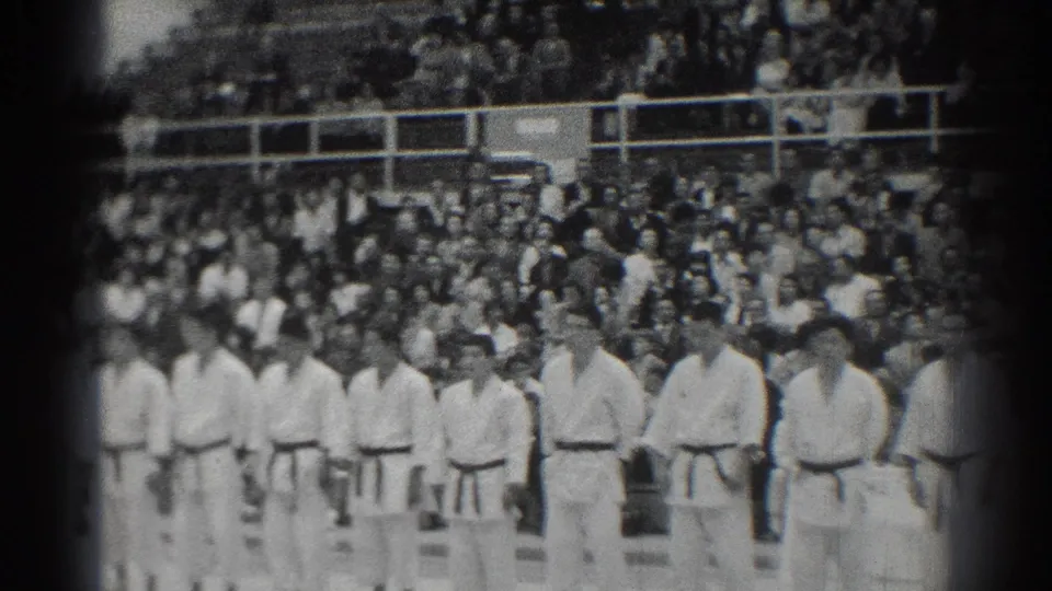 1966 San Gabriel frame of karate students in white gi standing in a gymnasium while spectators watch from bleachers.