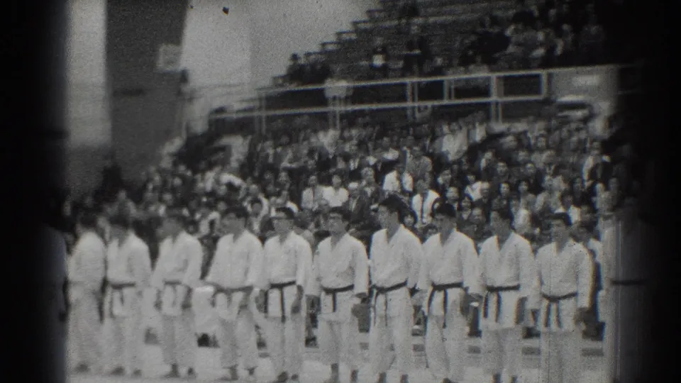 Judo athletes remain lined up in the 1966 San Gabriel venue, with a dense spectator section visible behind them.