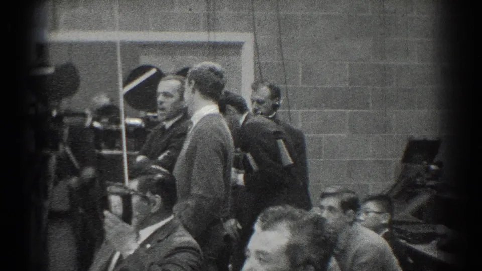 Sideline view from the 1966 San Gabriel judo event shows suited officials and photographers gathered beside the competition area.