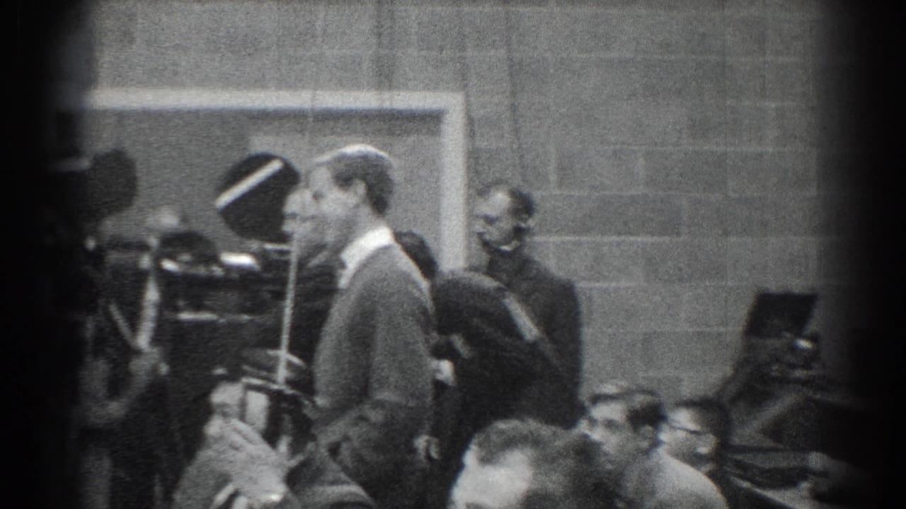 Black-and-white 1966 San Gabriel footage showing a suited man beside vintage cameras and seated observers in a formal room.