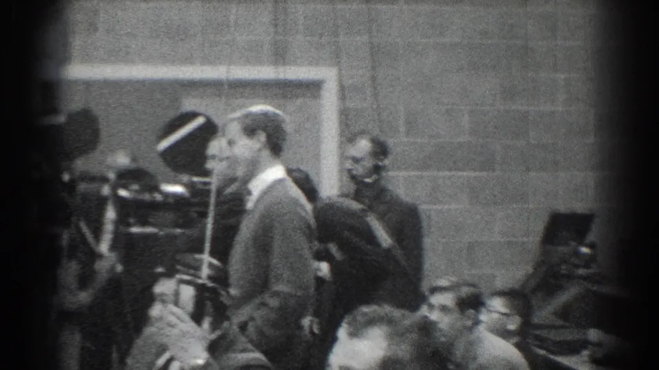 Black-and-white 1966 San Gabriel footage showing a suited man beside vintage cameras and seated observers in a formal room.