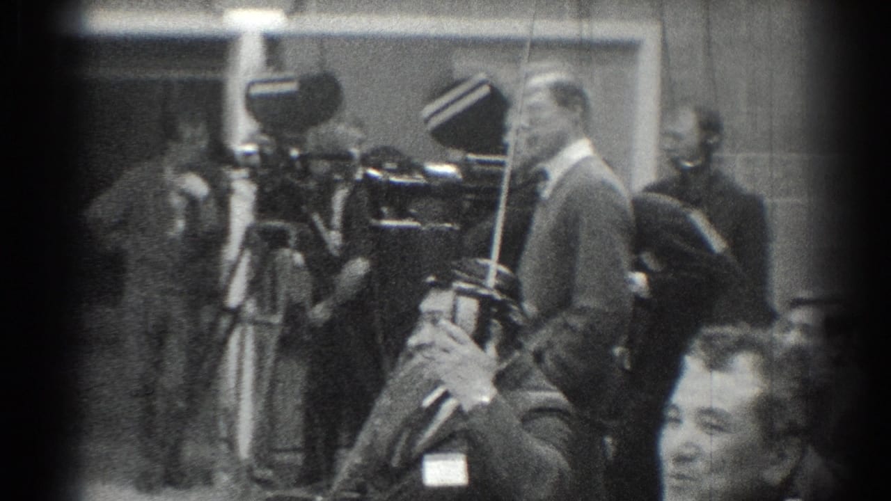 Camera operators and suited men stand near large tripods during an indoor event.