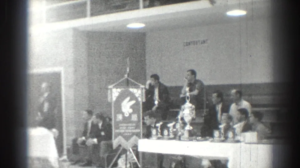 Black-and-white 8mm frame of a formal San Gabriel gathering with speakers, a banner, table objects, and seated attendees.