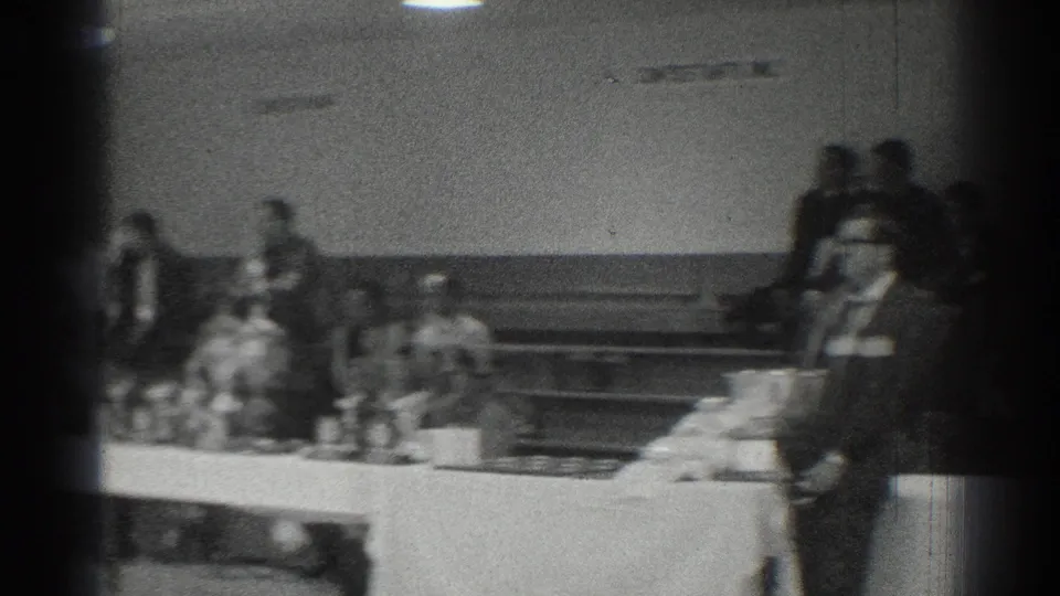 Wide black-and-white frame from San Gabriel showing seated observers and a speaker area in a cinder block interior.