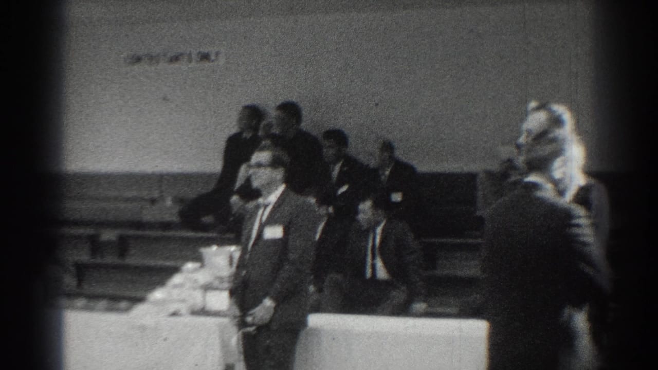 Men in suits stand near a table inside a gymnasium while spectators sit behind them.