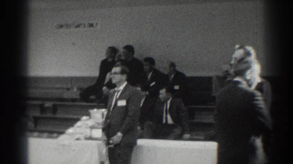 1966 San Gabriel school assembly frame showing suited adults with name tags near a table and bleachers in the background.
