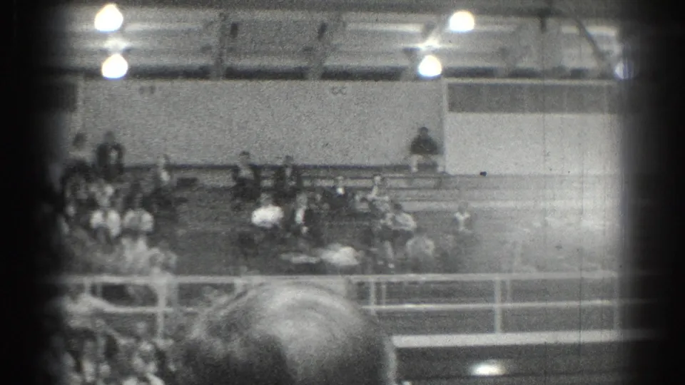 1966 San Gabriel home-movie frame showing gym bleachers with spectators, overhead lights, railings, and heavy 8mm edge shadowing.