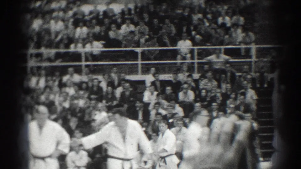 1966 San Gabriel frame showing martial arts students or instructors in white gi near a large seated audience in gym bleachers.
